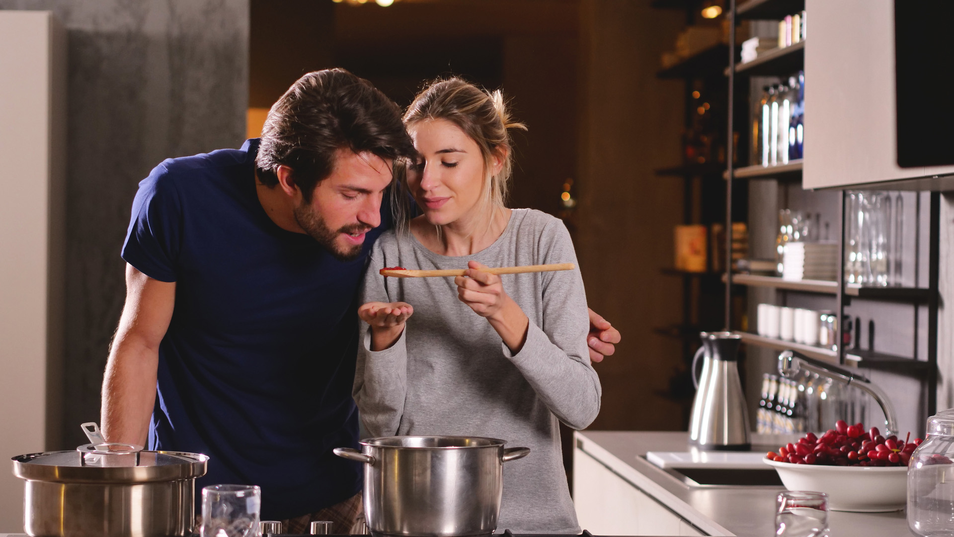 Brunette man in navy t-shirt being fed red sauce from cooking spoon by blonde woman in grey t-shirt. They are in the kitchen.