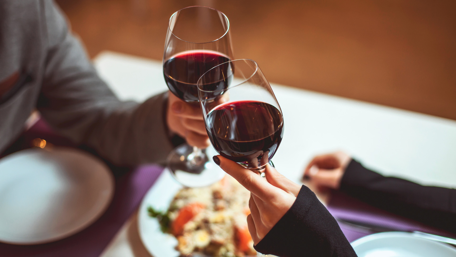 Man and woman making a toast with their wine glasses which have red wine. They are sitting down on a table and having dinner.