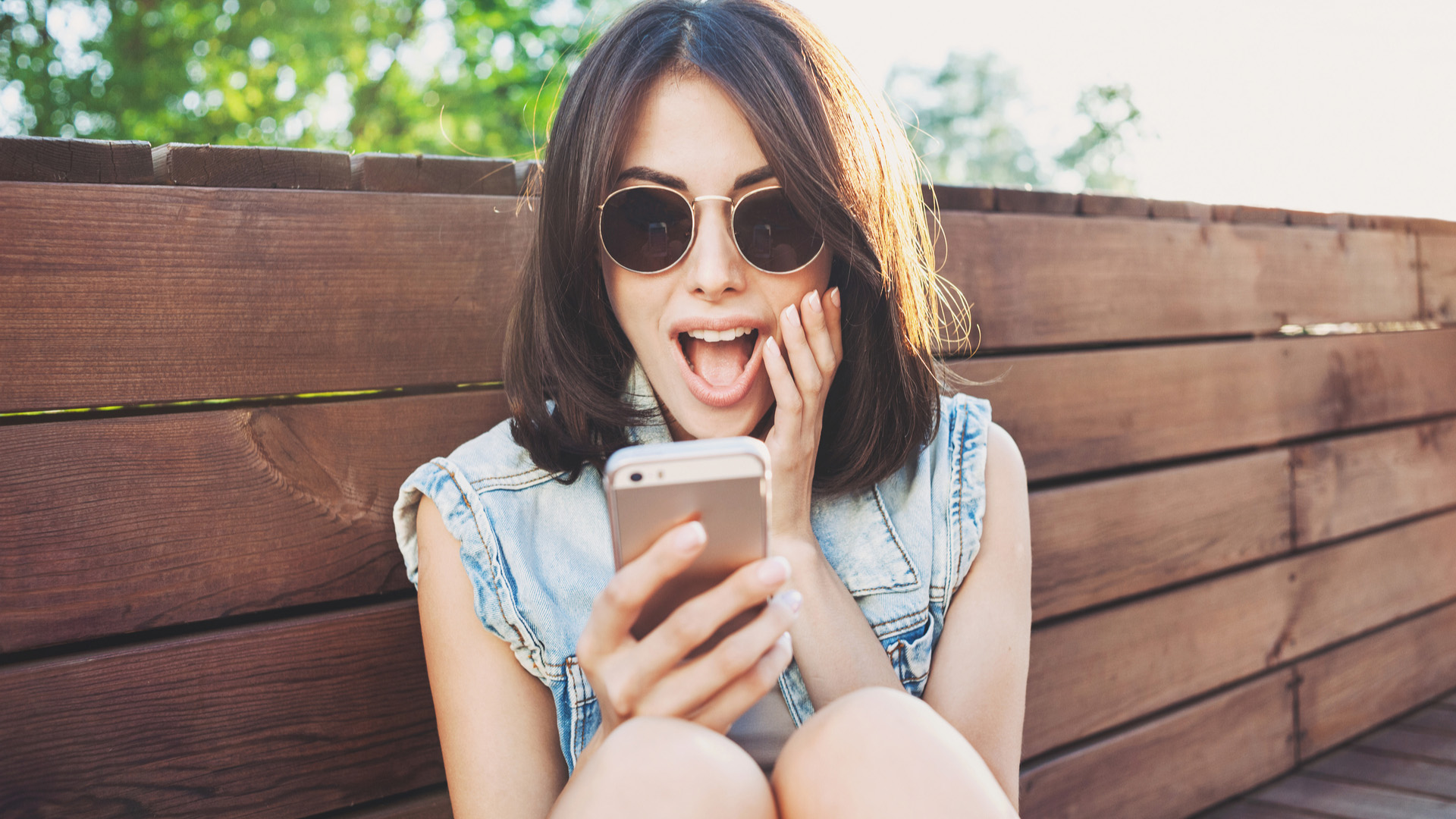 Brunette woman in sunglasses and jean sleeveless jacket, sitting on wooden bench, looking surprised at her mobile phone.