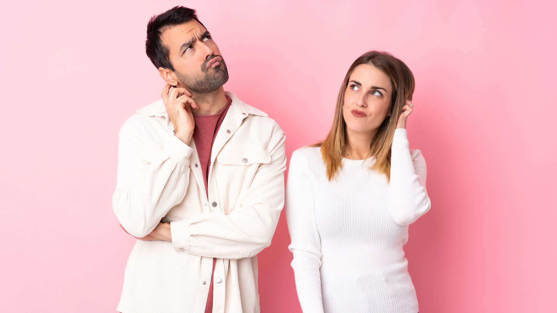 Brunette man and blonde woman both wearing white, scratching their heads and looking up in question. Pink background.