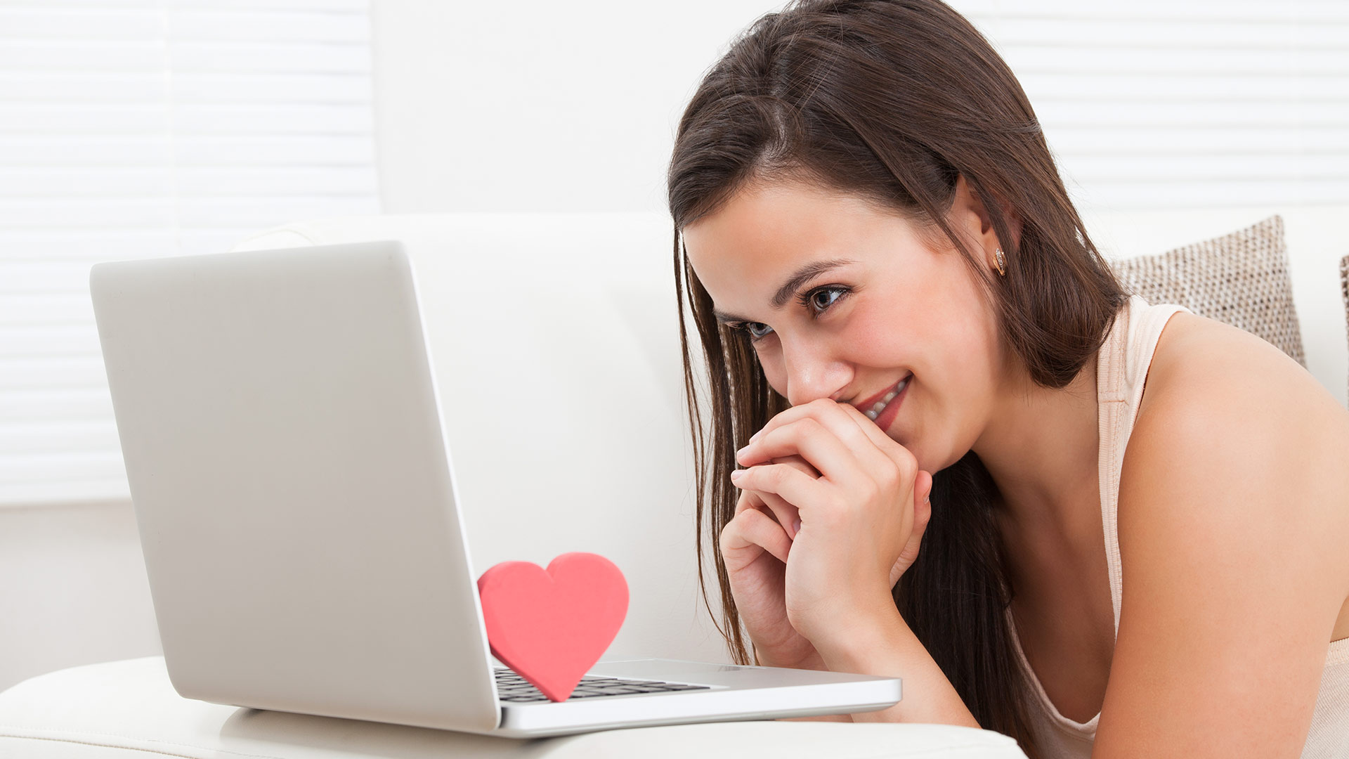 Brunette woman wearing a white tank top smiling while looking at her laptop screen. There is a red love heart on the keyboard