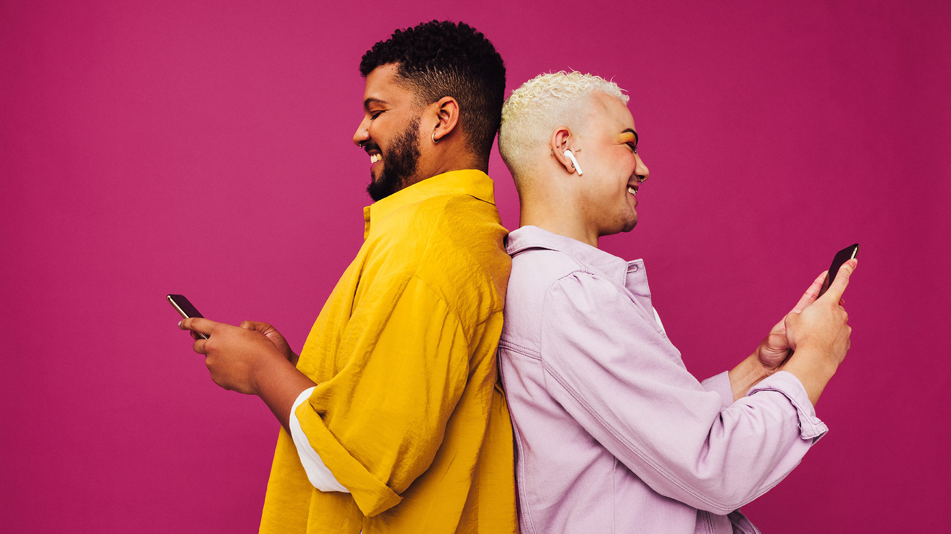 Brunette man in yellow shirt and blonde man in pink shirt texting while leaning against each other and smiling.