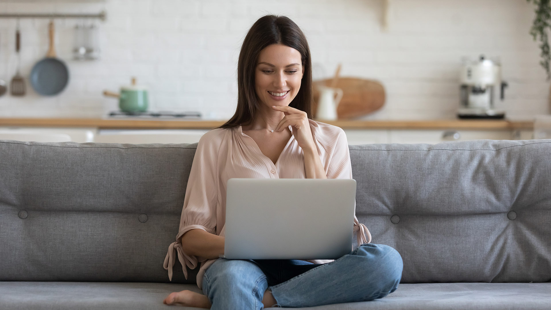 Brunette woman smiling while looking at her laptop screen. She is sitting on a grey couch with a kitchen counter behind it.