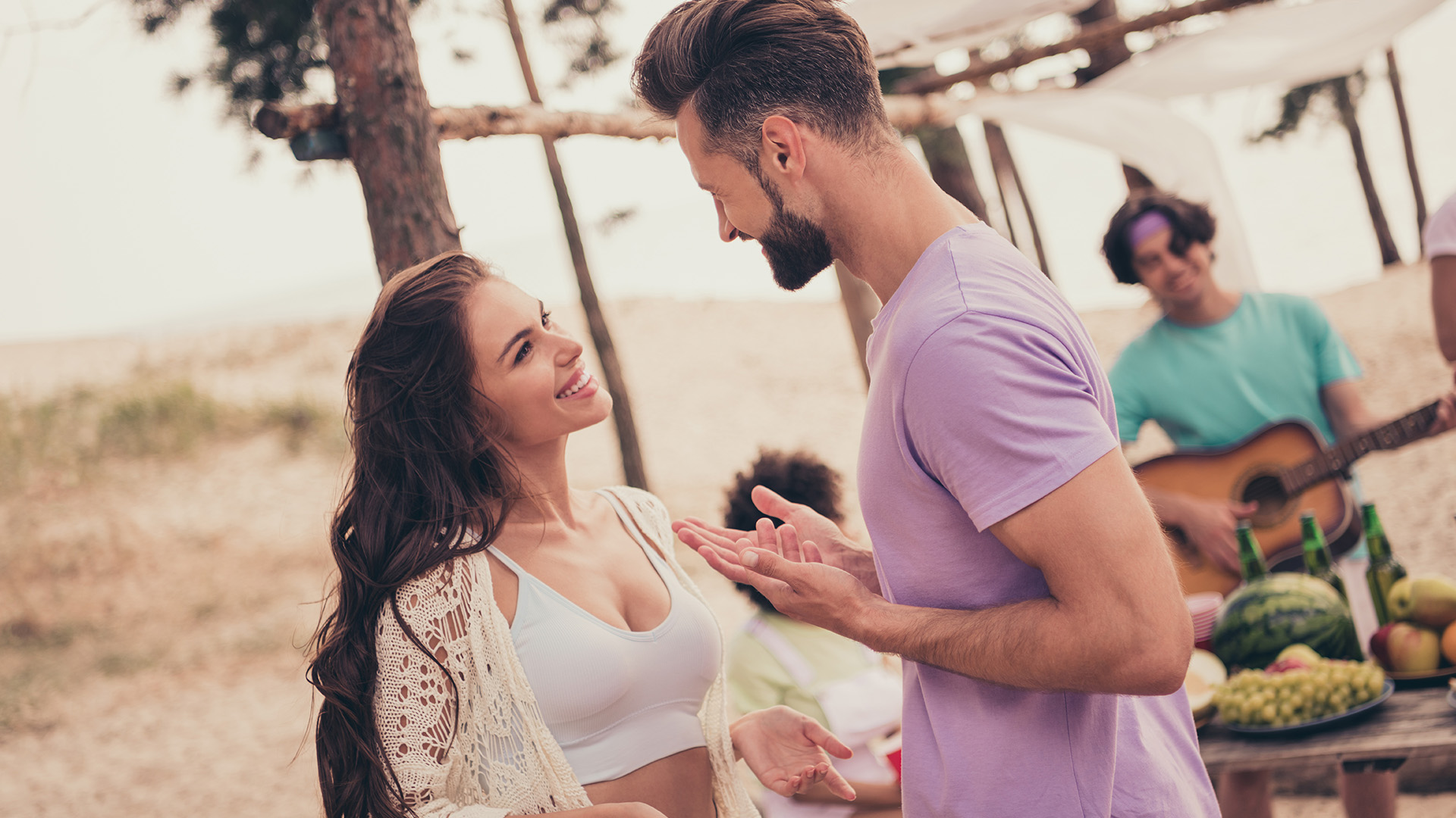 Brunette man in pink shirt and woman in white bustier smiling at each other while talking. Beach in the background.