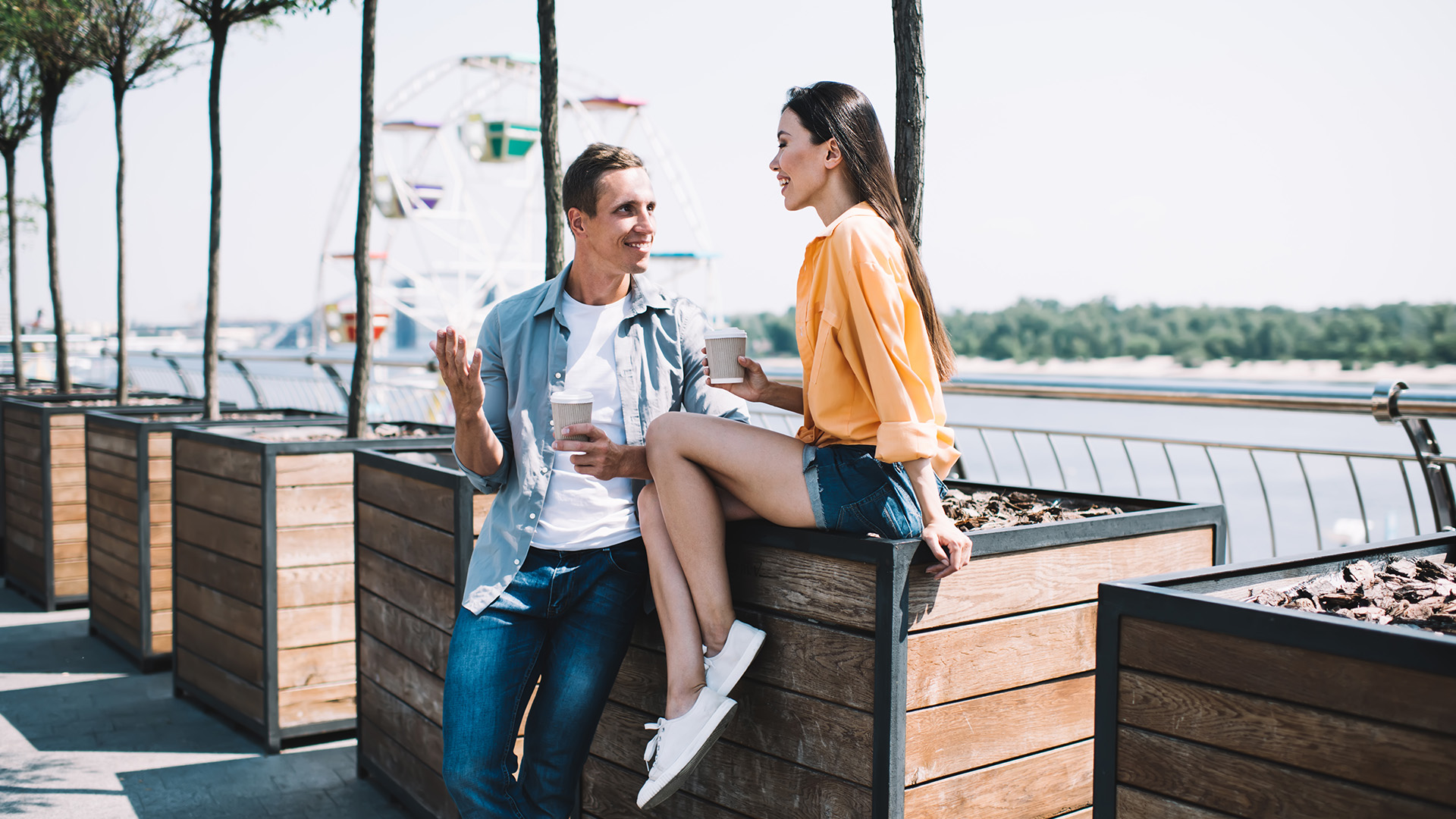 Man standing next to a woman, who is sitting on a wooden pot next to him chatting and drinking coffee near a river.