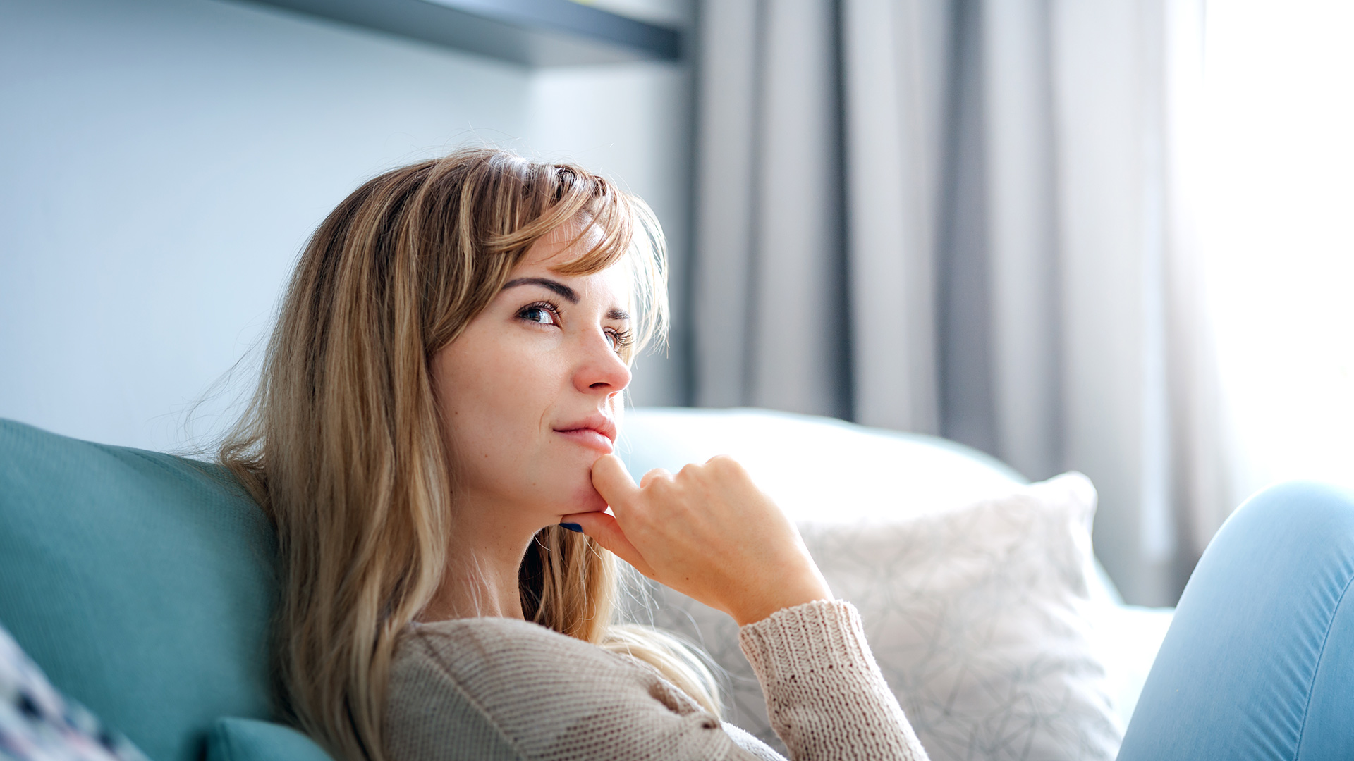 Woman with fringe bangs, wearing a grey sweater and blue jean, sitting on a turquoise couch, rubbing her chin while thinking.