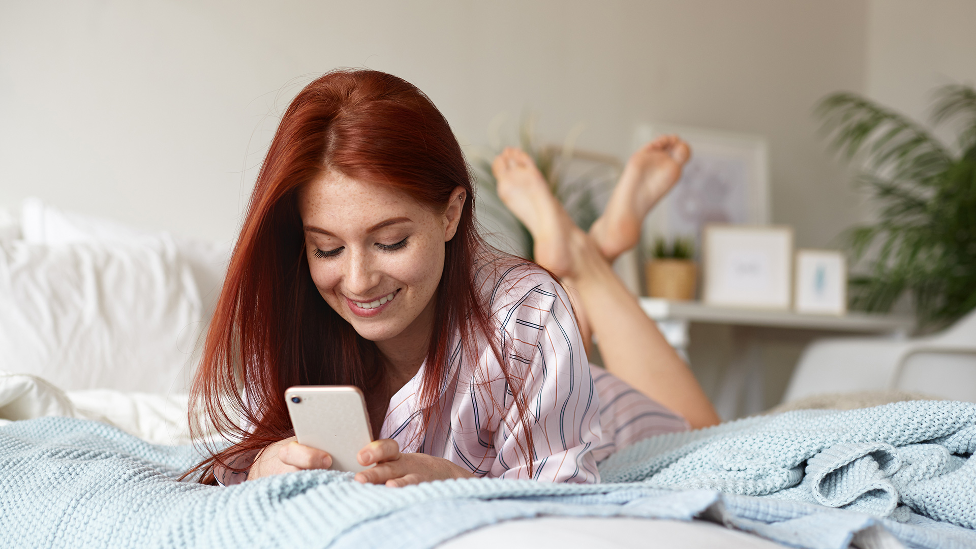 Red-haired woman in her pajamas lying in bed while crossing her legs behind her. She is looking at her phone and smiling.