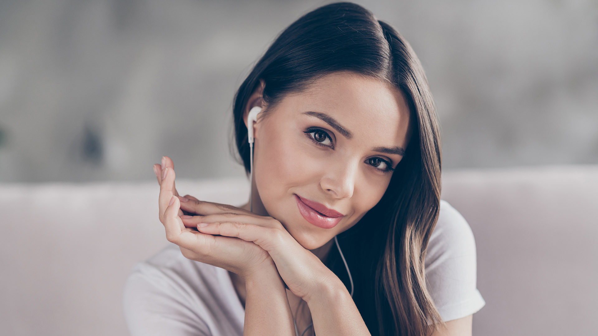 Brunette woman in white shirt, wearing white headphones and resting her head on her hands. She is smiling.