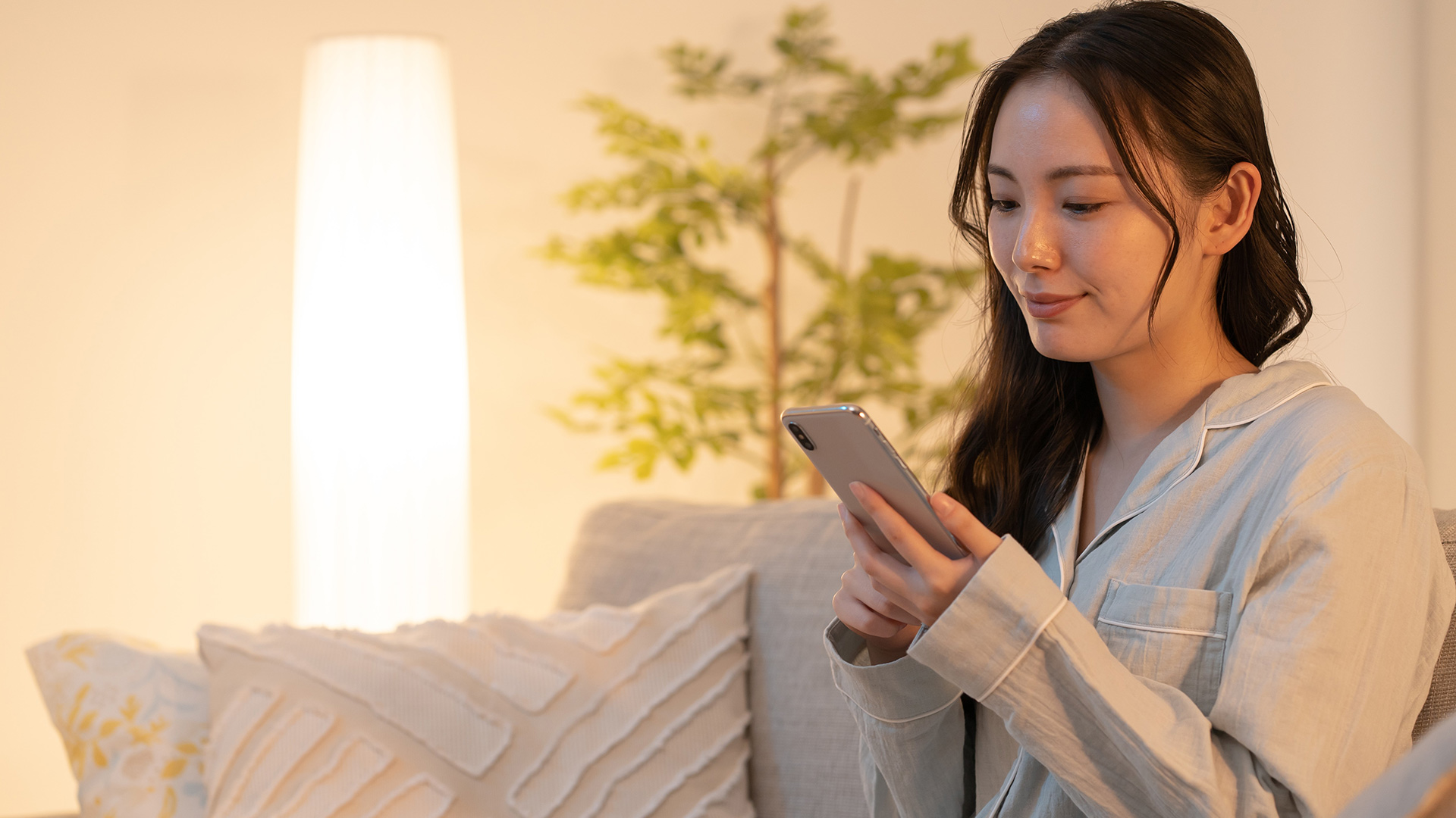 Brunette woman in beige pajamas, sitting down on a grey couch and holding her phone. She is thinking about something.
