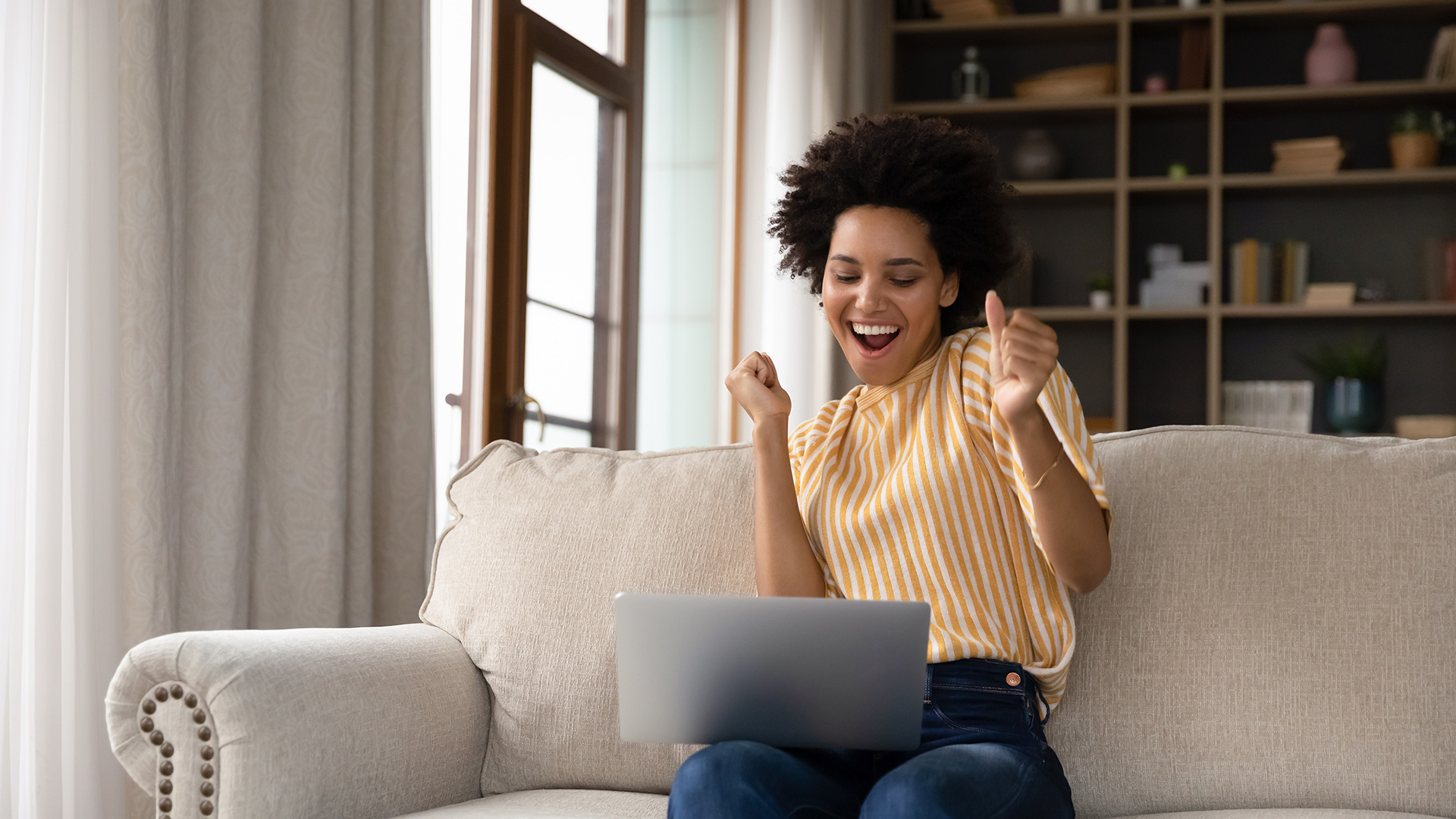 Woman with black hair, wearing a yellow-white striped shirt, sitting down and dancing happily in front of laptop on her lap.