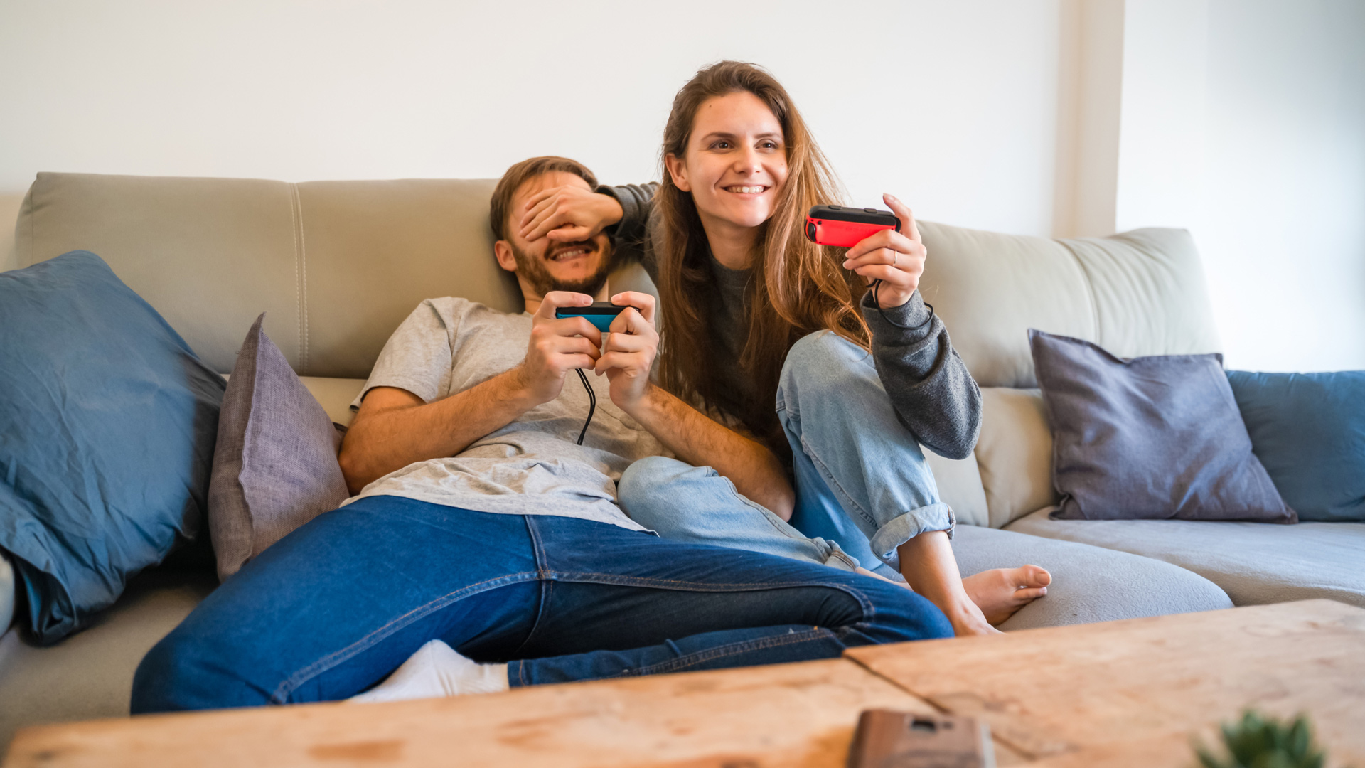 Man and woman sitting on a couch. They are both holding controllers and the woman is covering the man's eyes with her hand.