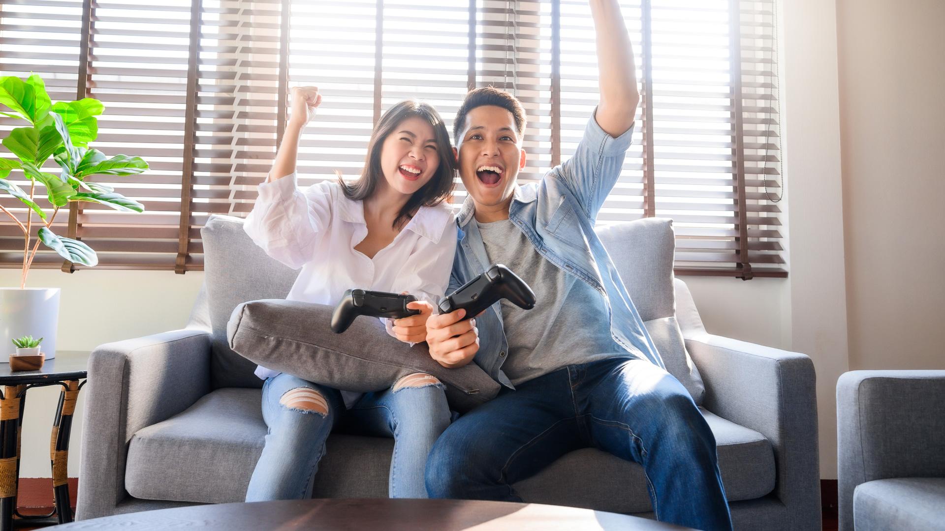 Man and woman sitting on a couch, both holding a controller and raising their arms in celebration.