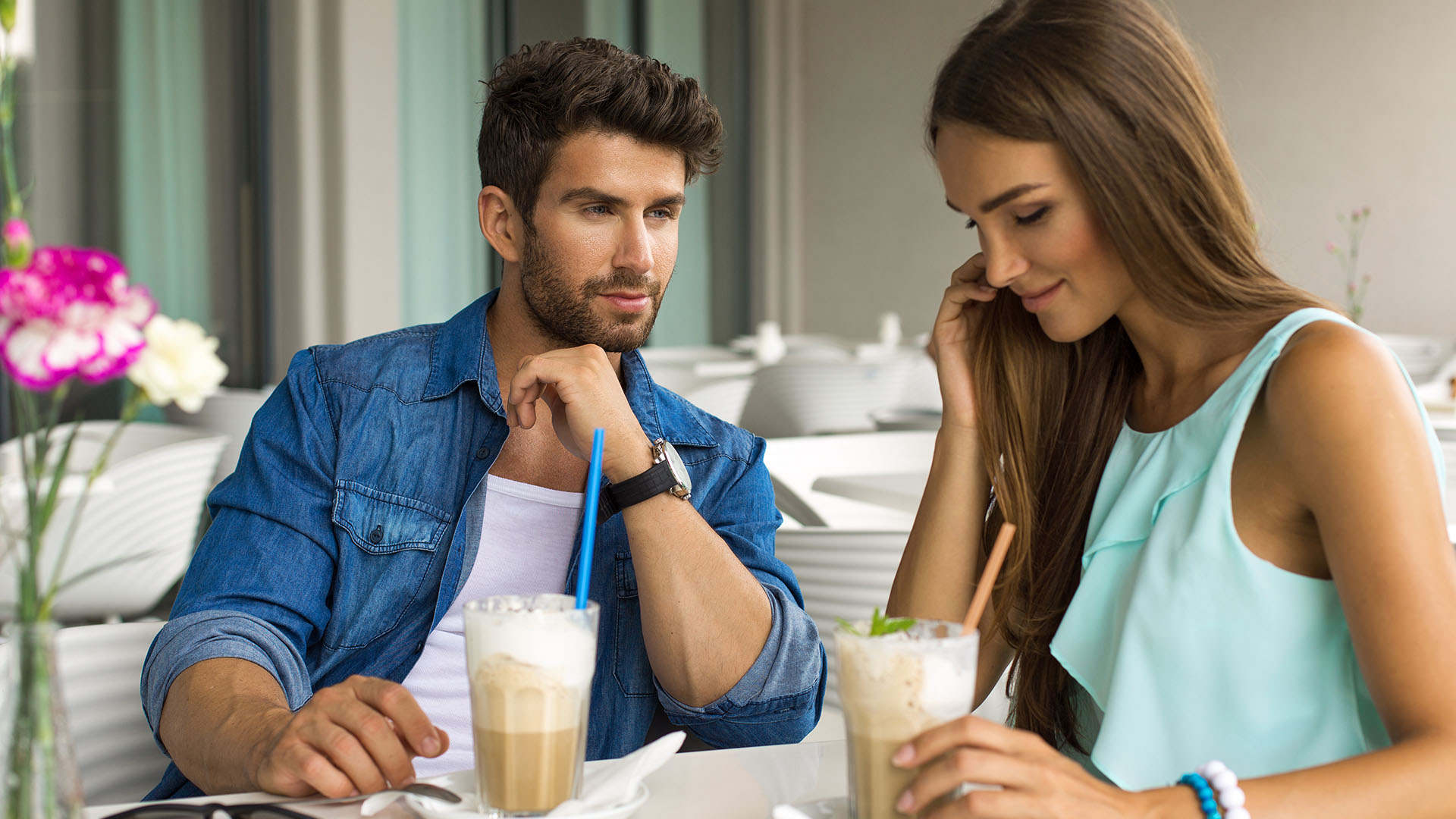 Man in blue-jean shirt sneering at brunette woman sitting next to him and having a coffee. She is smiling while looking down.