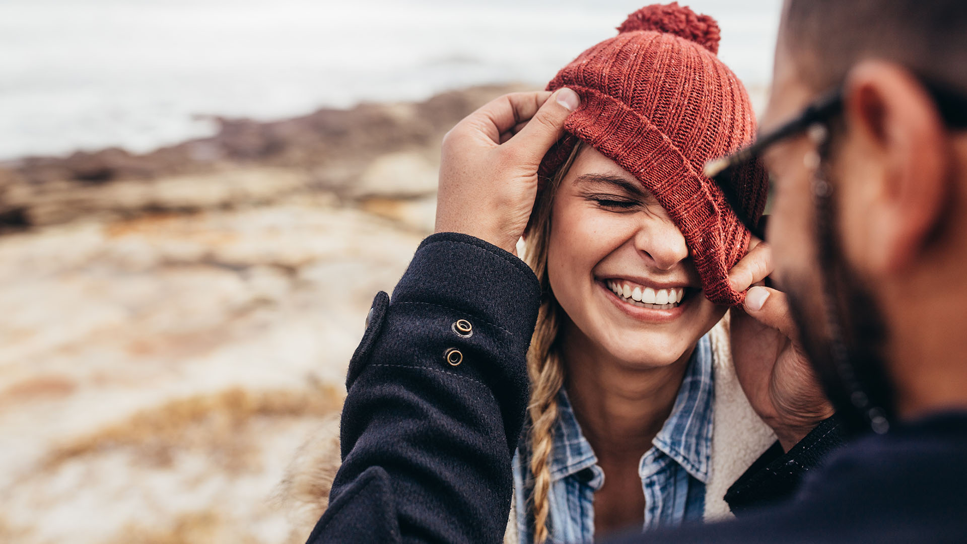 Blonde woman laughing while brunette man in sunglasses crooks red beanie over her face. Ocean in the background.