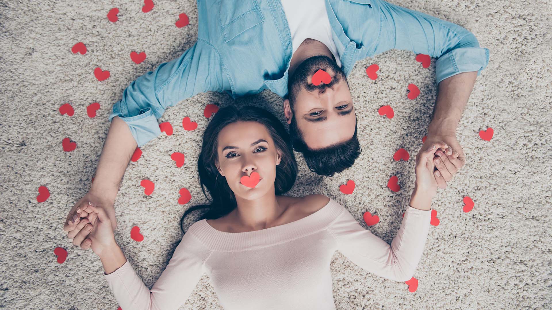 Brunette man in blue shirt and woman in white shirt lying on beige carpet with love hearts on their lips, holding hands.
