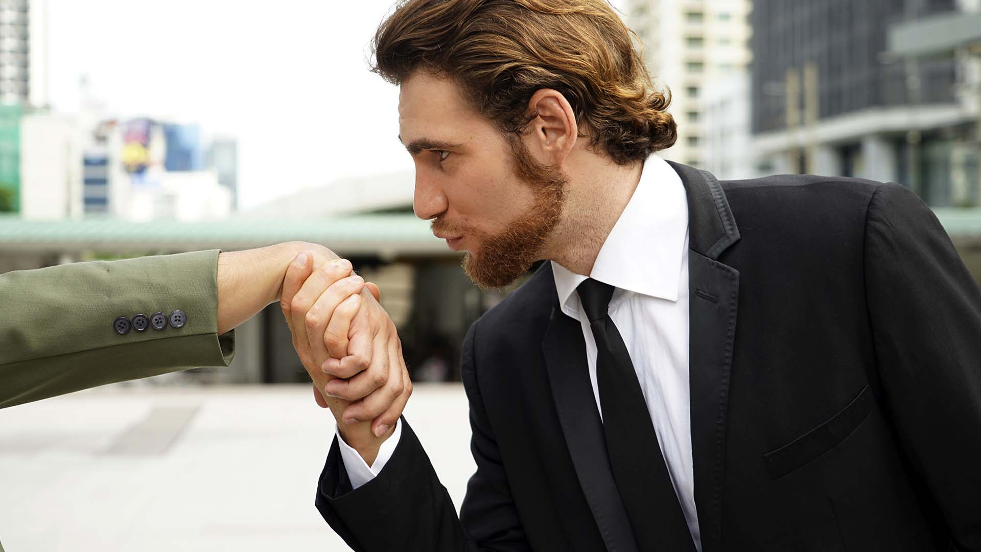 Blonde bearded man wearing a black tuxedo holding a woman's hand and leaning closer to kiss it while looking up towards her.