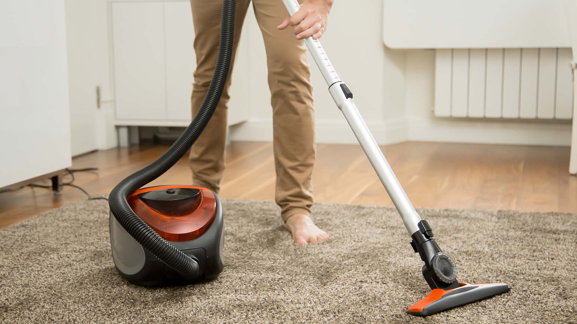 Lower body of a man wearing beige trousers holding a hoover and using it on a carpet. He is barefoot on the carpet.
