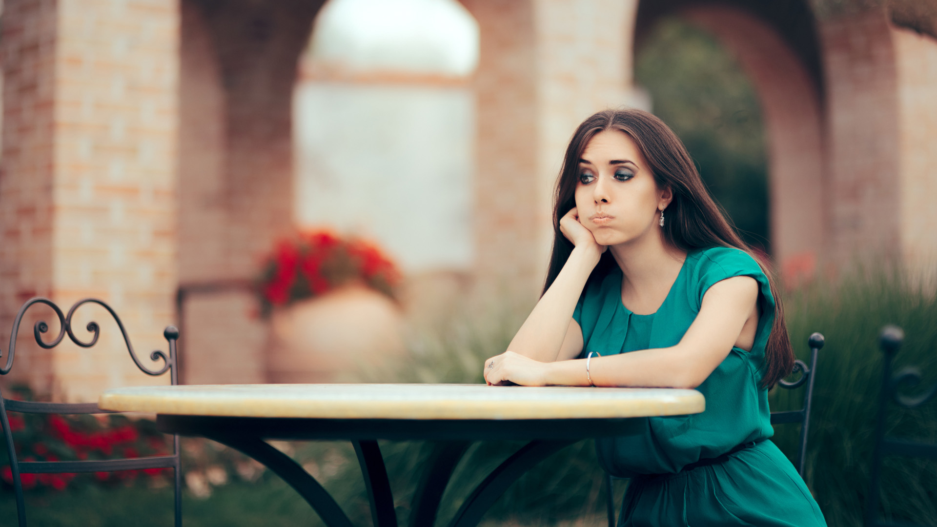 Brunette woman in turquoise dress sitting alone at a circular table resting her head in her palm and pouting her lips.