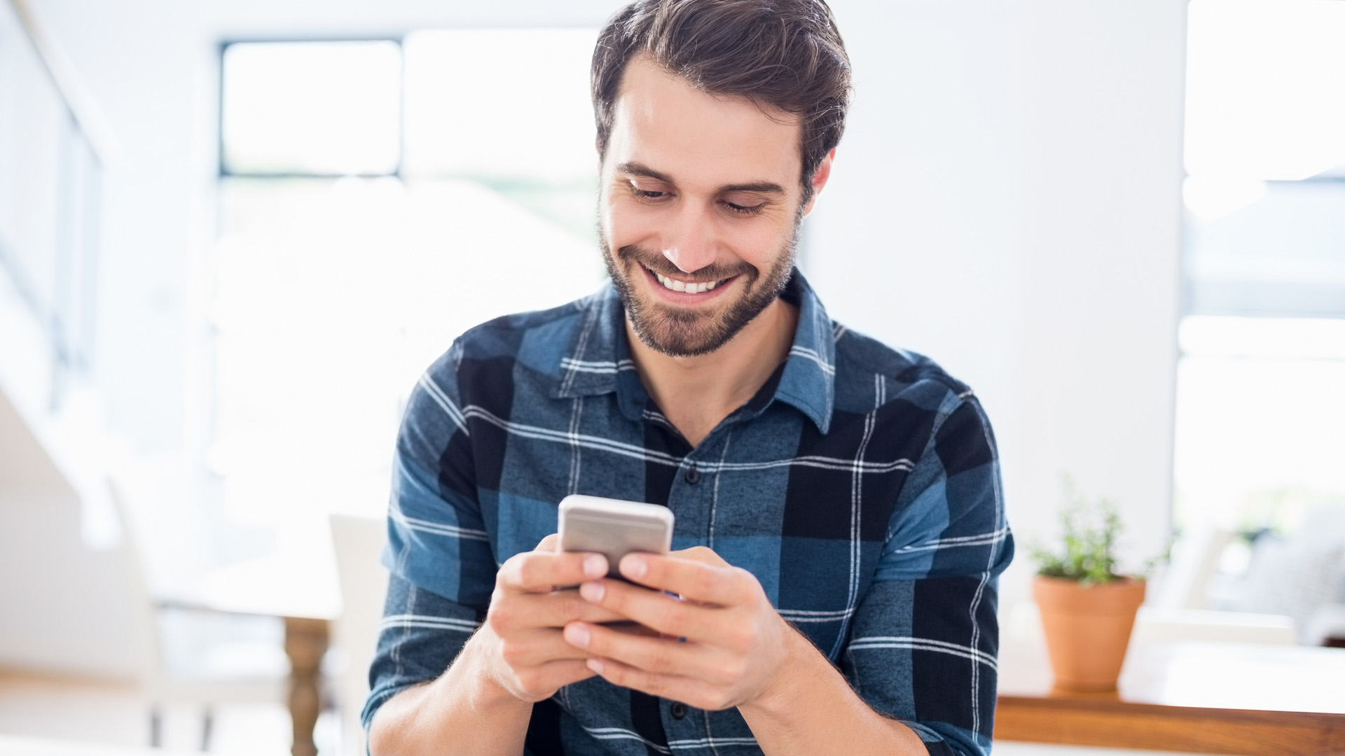 Brunette man in blue checkered shirt holding his phone with both hands and smiling while looking at it. Background flower pot