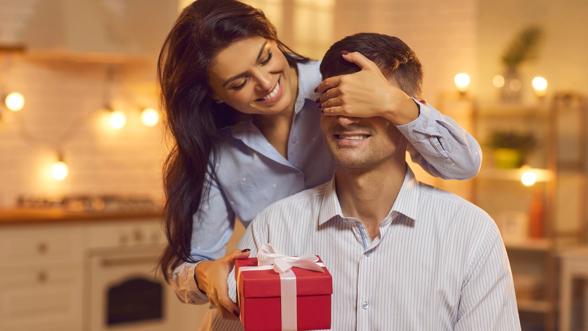 Brunette woman covering eyes of man with one hand and placing red gift box in front of him. Light bulbs in background.