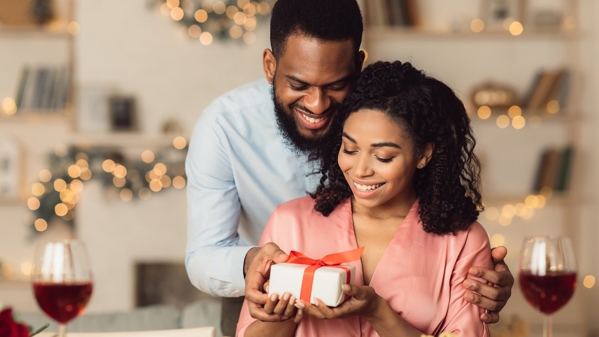 Man in blue shirt hugging a woman in pink shirt from behind while giving her a gift box. Two red wine glasses in front.