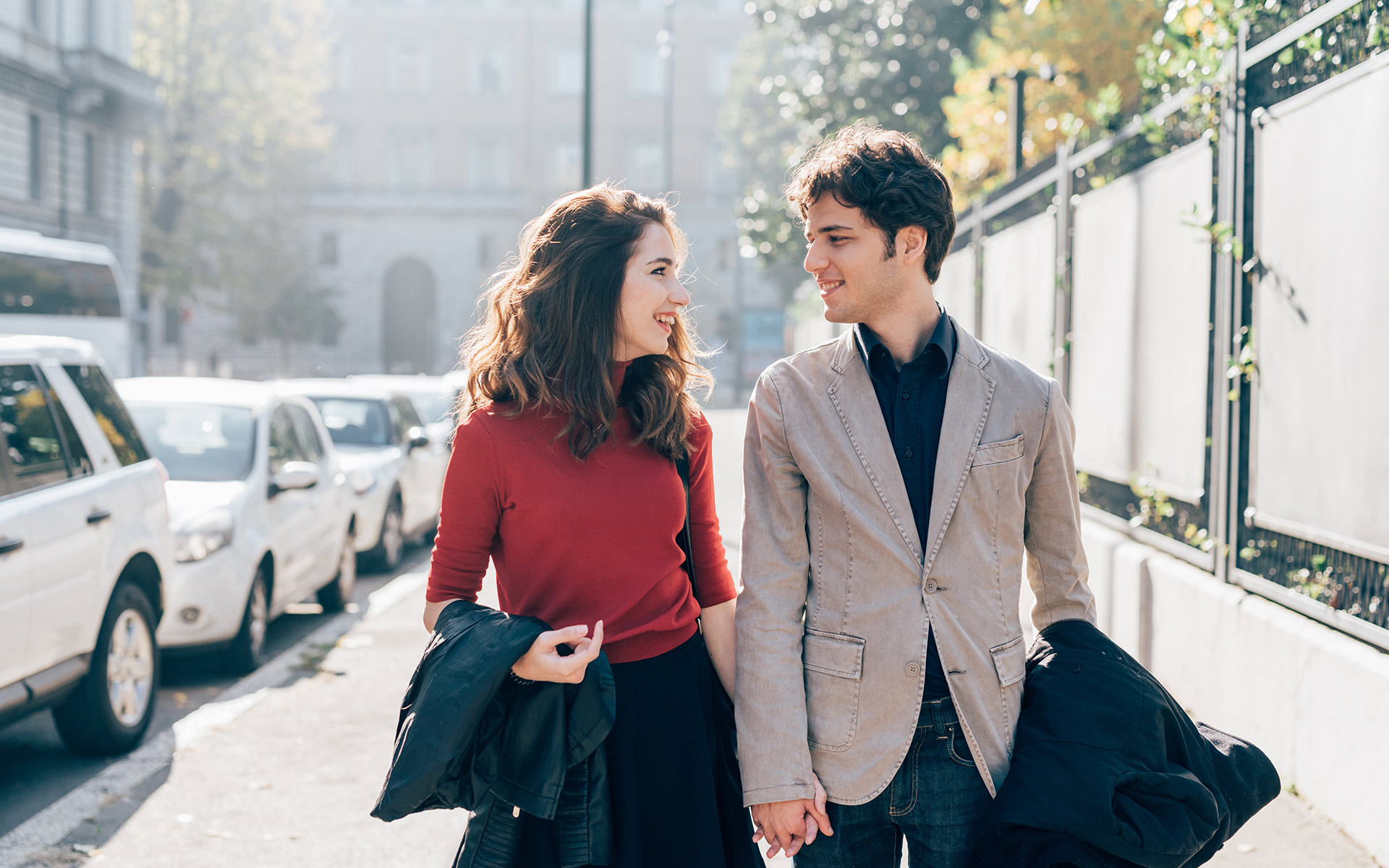 Brunette man in grey coat and brunette woman in red jumper holding hands, walking down the road and looking at each other.