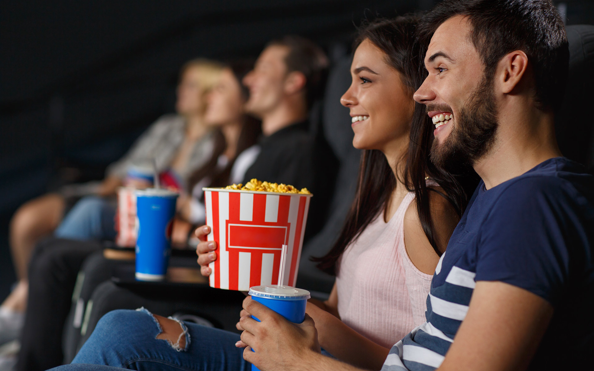 Brunette man and woman on a date at the cinema theatre. The man is holding a drink and the woman a pack of popcorn.