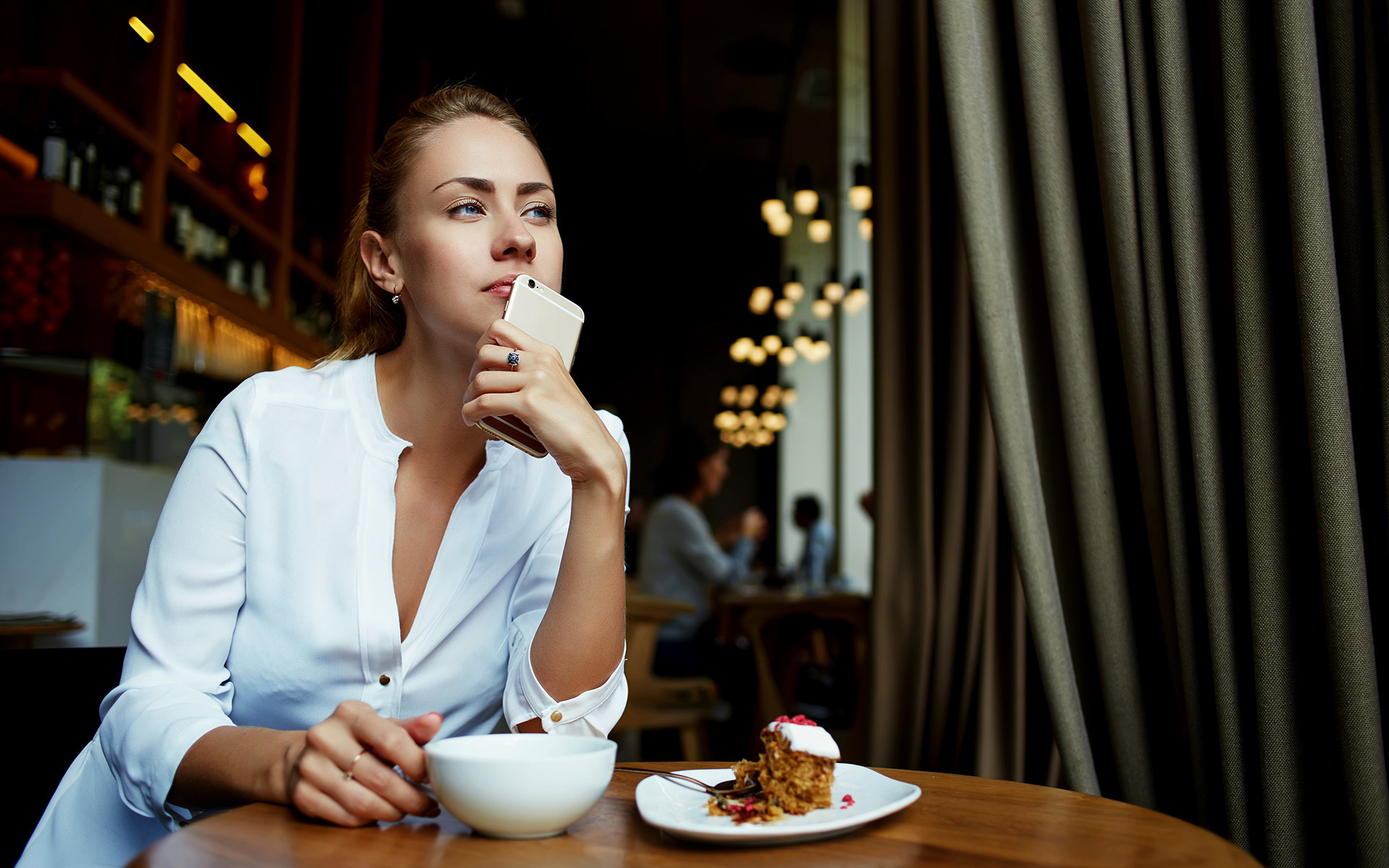 Blonde woman in white shirt holding her phone up to her lips while thinking. She is having a coffee and cake at a cafe.