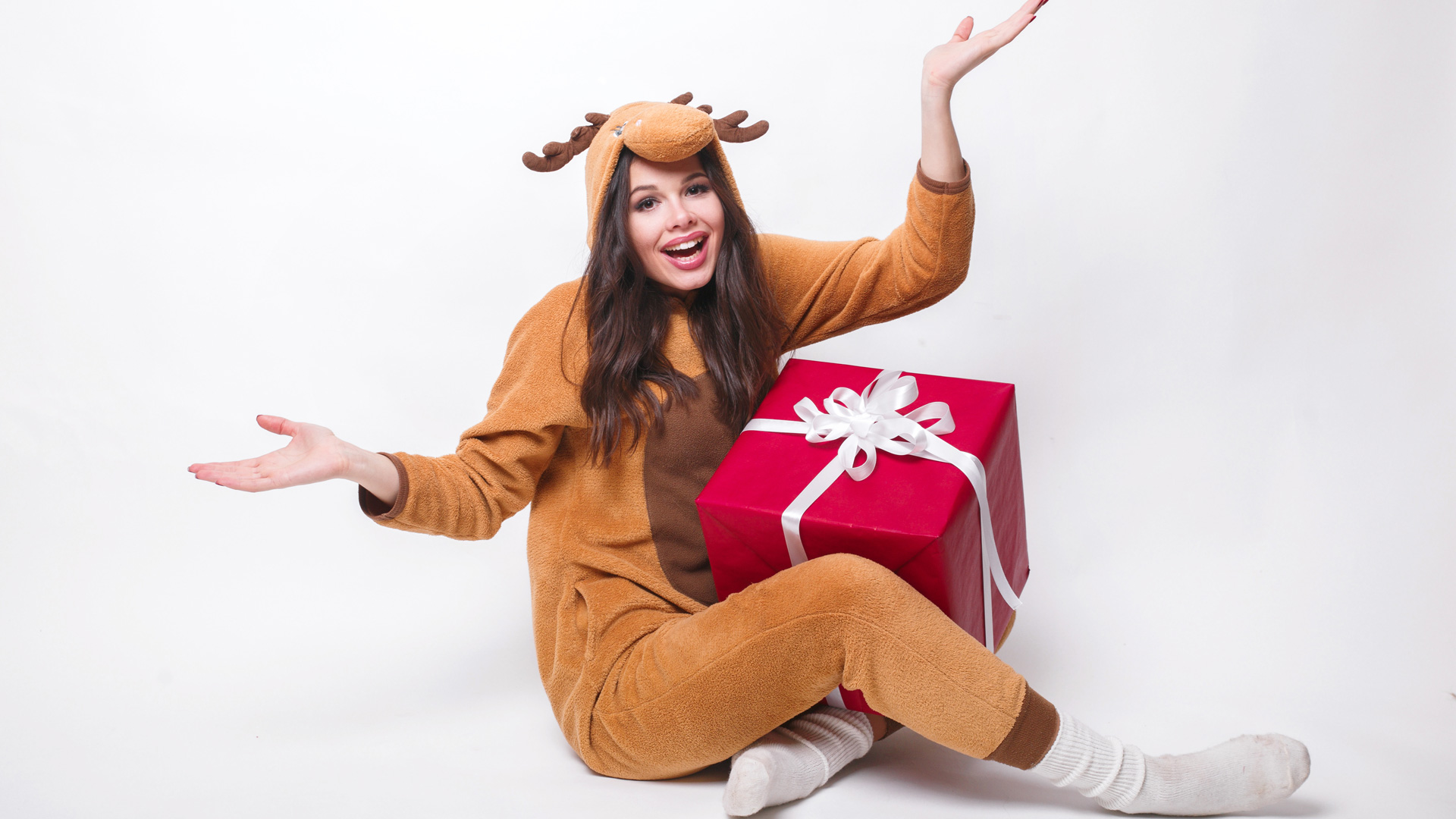 Brunette woman with long hair sitting down with red gift in her lap dressed in a brown reindeer onesie.