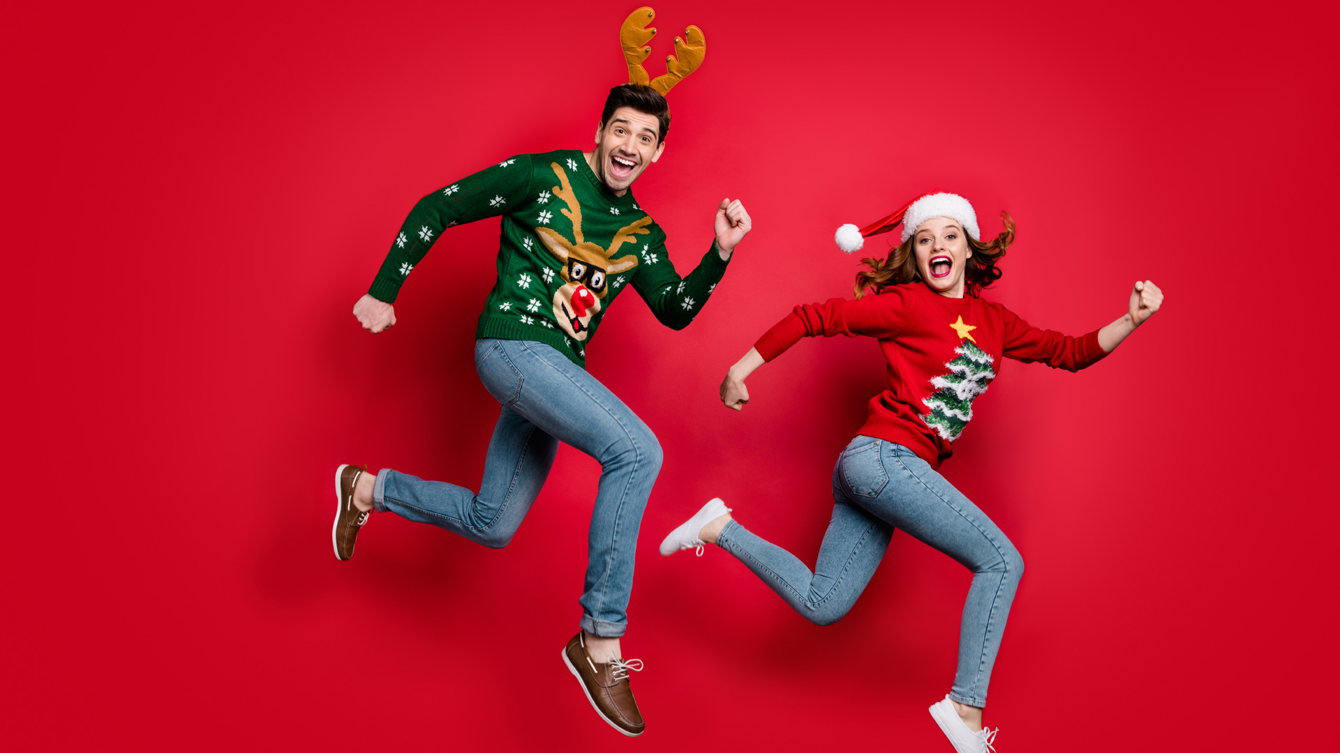 Couple dressed in green and red Christmas jumpers with reindeer and tree on them, wearing jeans and jumping in the air.
