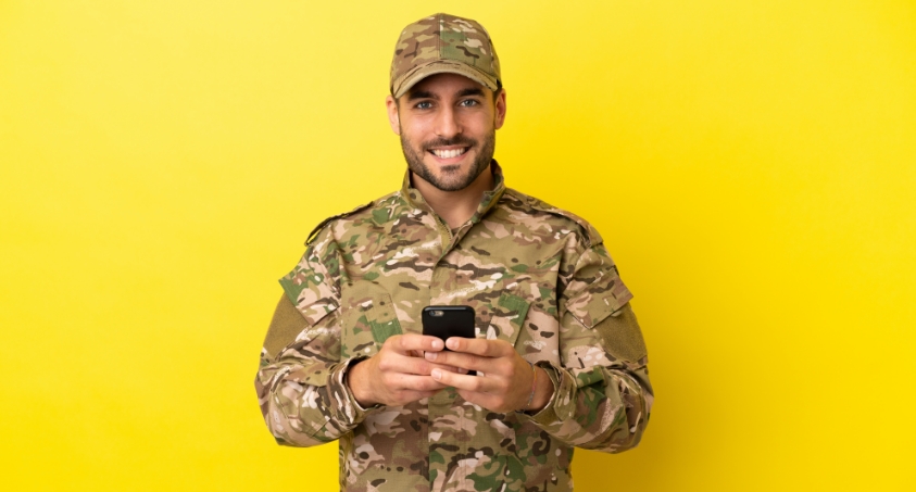 Forces penpals man in army attire holding his mobile phone with both hands and smiling. Yellow background.