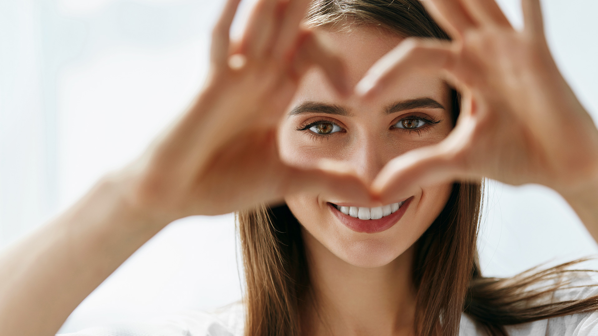 Brunette woman forming the shape of a love heart with her fingers in front of her face, framing her eyes. She is smiling.