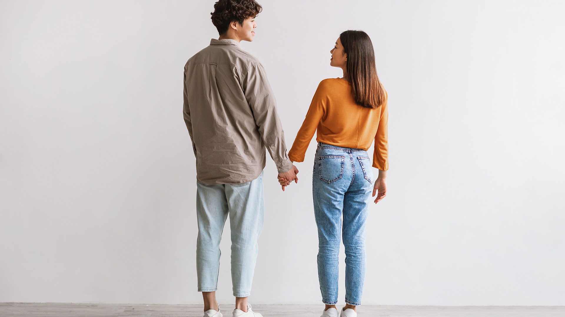 Back of brunette man in beige shirt and brunette woman in yellow shirt, holding hands and looking at each other.