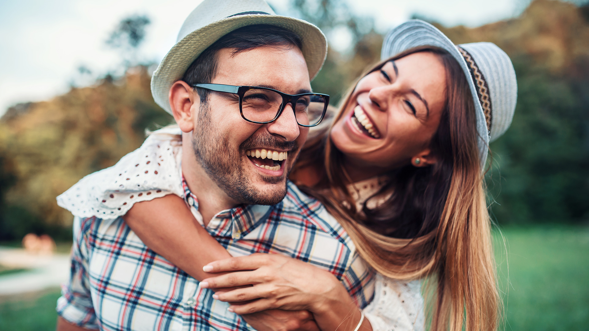 Man and woman wearing white straw hats, laughing together. The woman is hugging the man from behind.