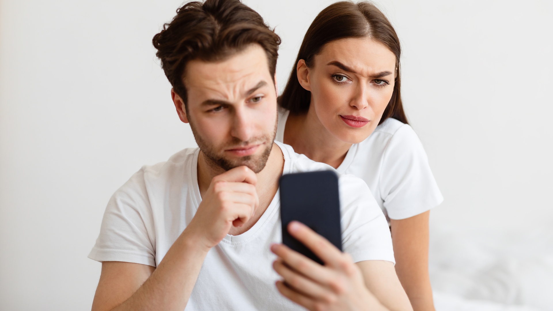 Brunette man and woman wearing white shirts. The man is looking at his phone screen while the woman is peaking behind him.