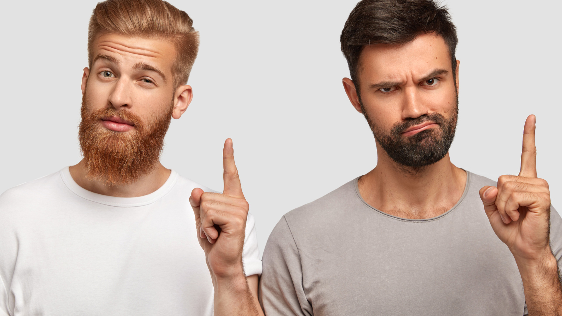 Brunette man with grey t-shirt and blonde man with white shirt both looking perplexed while holding their index fingers up.