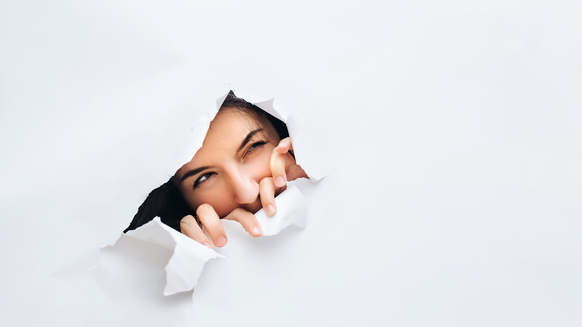 Face of a brunette woman looking suspicious coming out of a torn white paper wall.