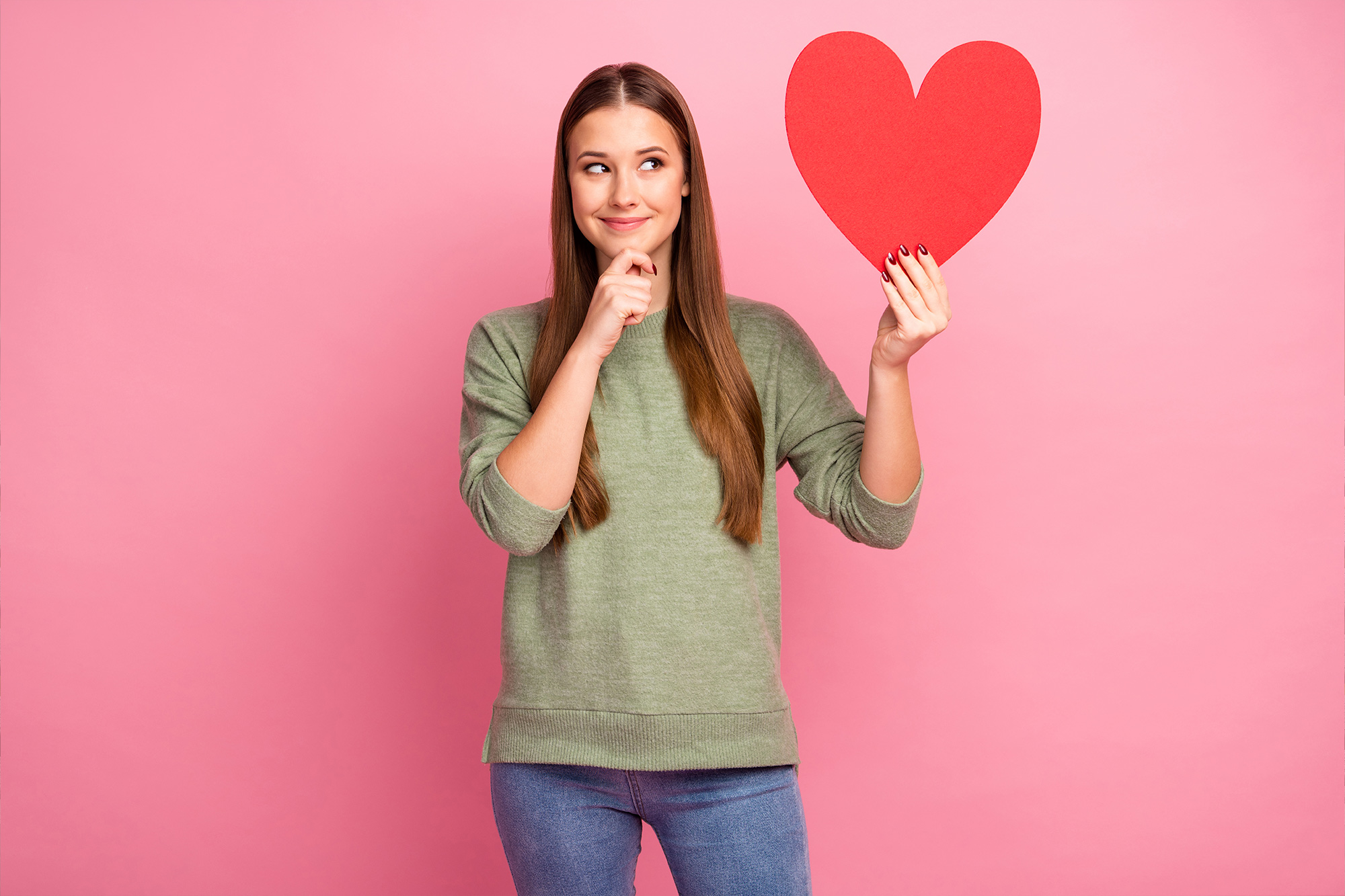 Brunette woman in green sweater and blue jeans holding a red love heart with one hand and her chin with the other.