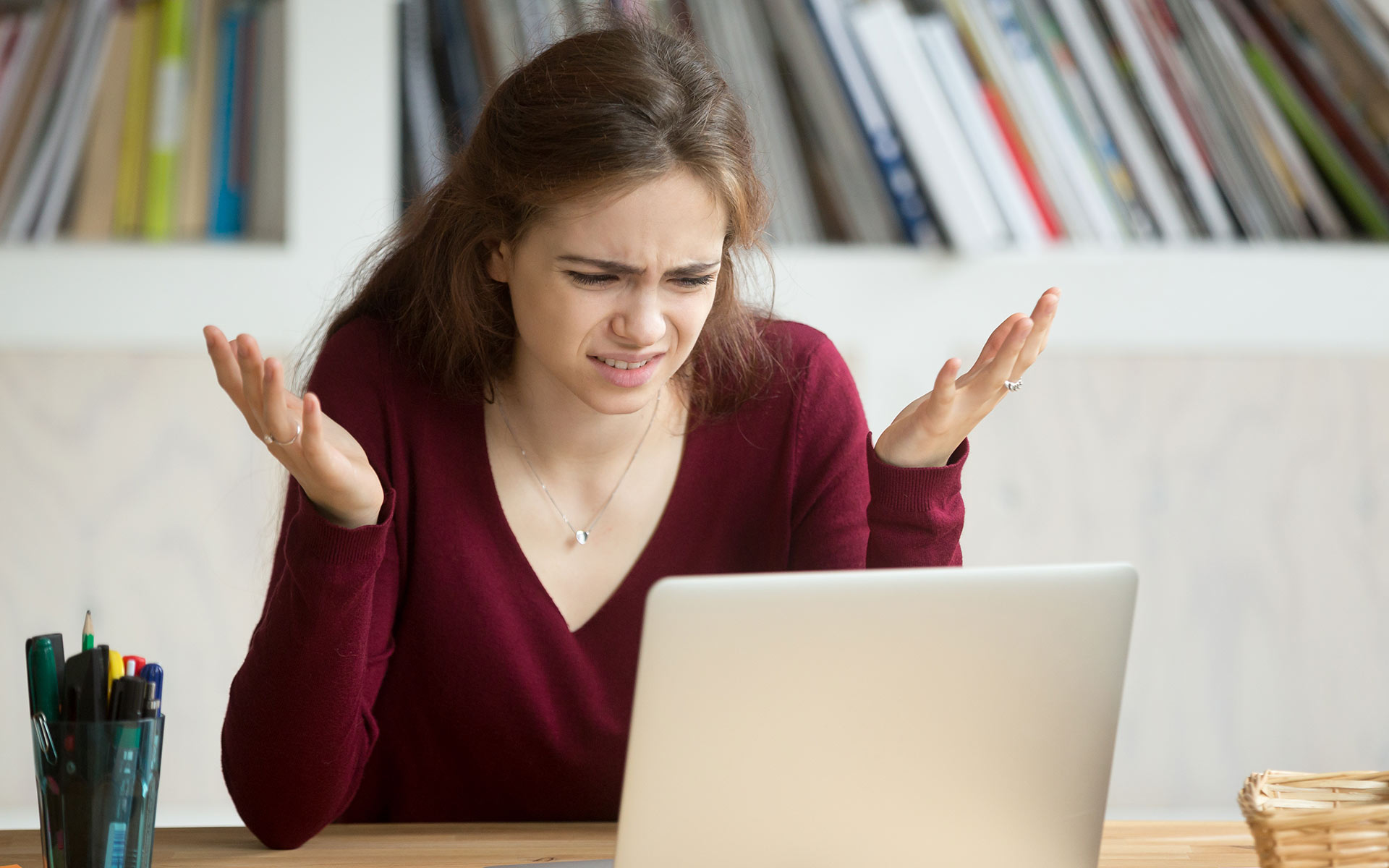 Brunette woman in dark red sweater holding her arms up in a shrugging motion while looking angrily at her laptop.