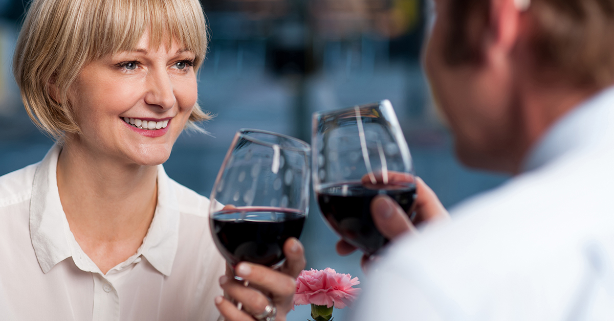 Elderly man and elderly blonde woman with wine glasses containing red wine making a toast.