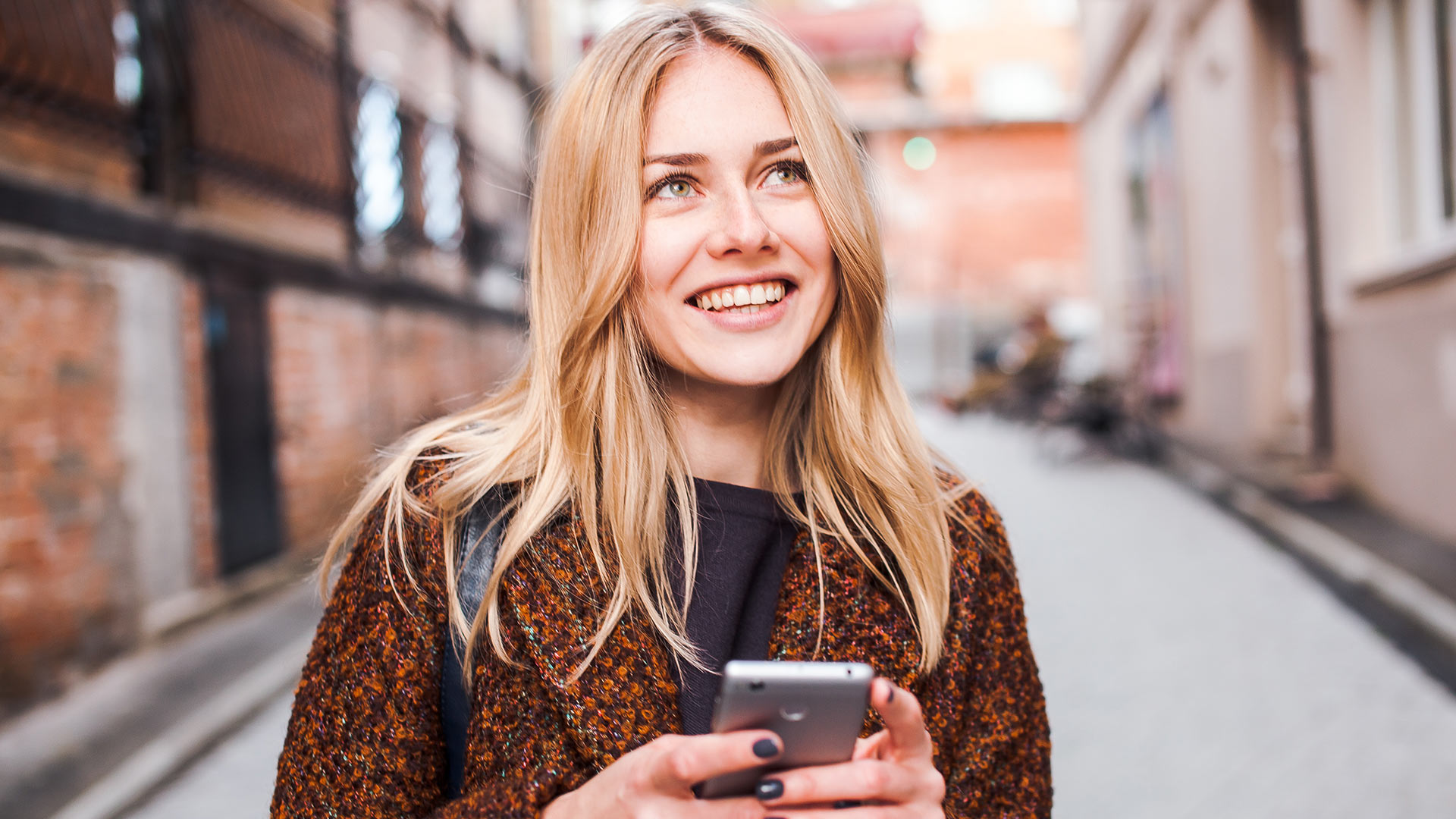 Blonde woman in black and orange coat holding her phone with both hands and smiling while looking up.