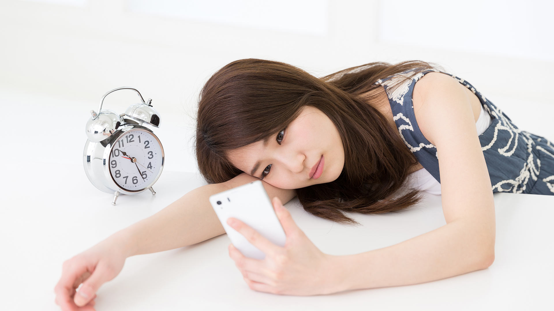 Brunette woman lying on a white desk with an alarm clock next to her, looking at her phone expectantly.