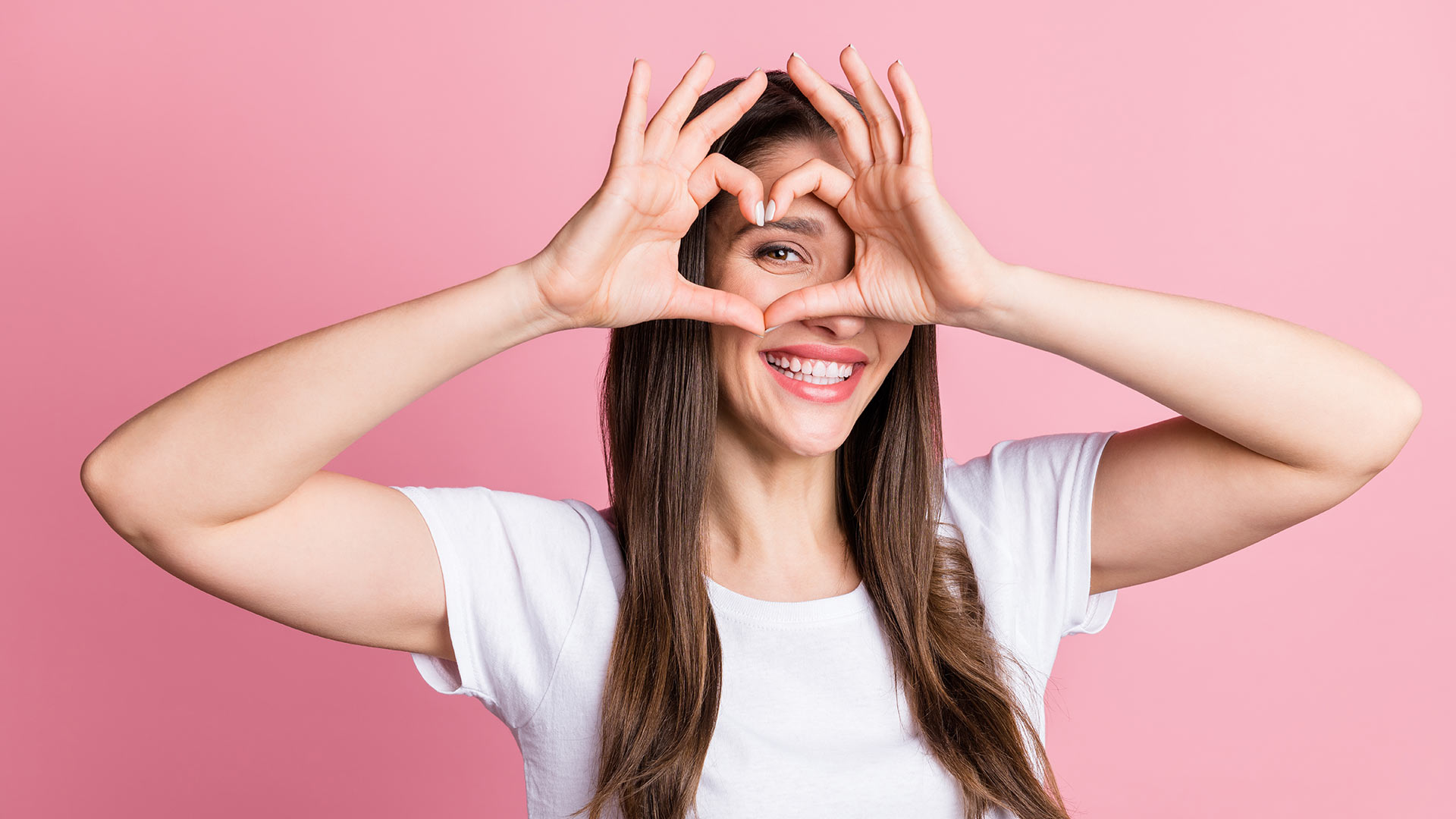 Brunette woman in white t-shirt smiling while making the shape of a love heart with her fingers in front of pink background.
