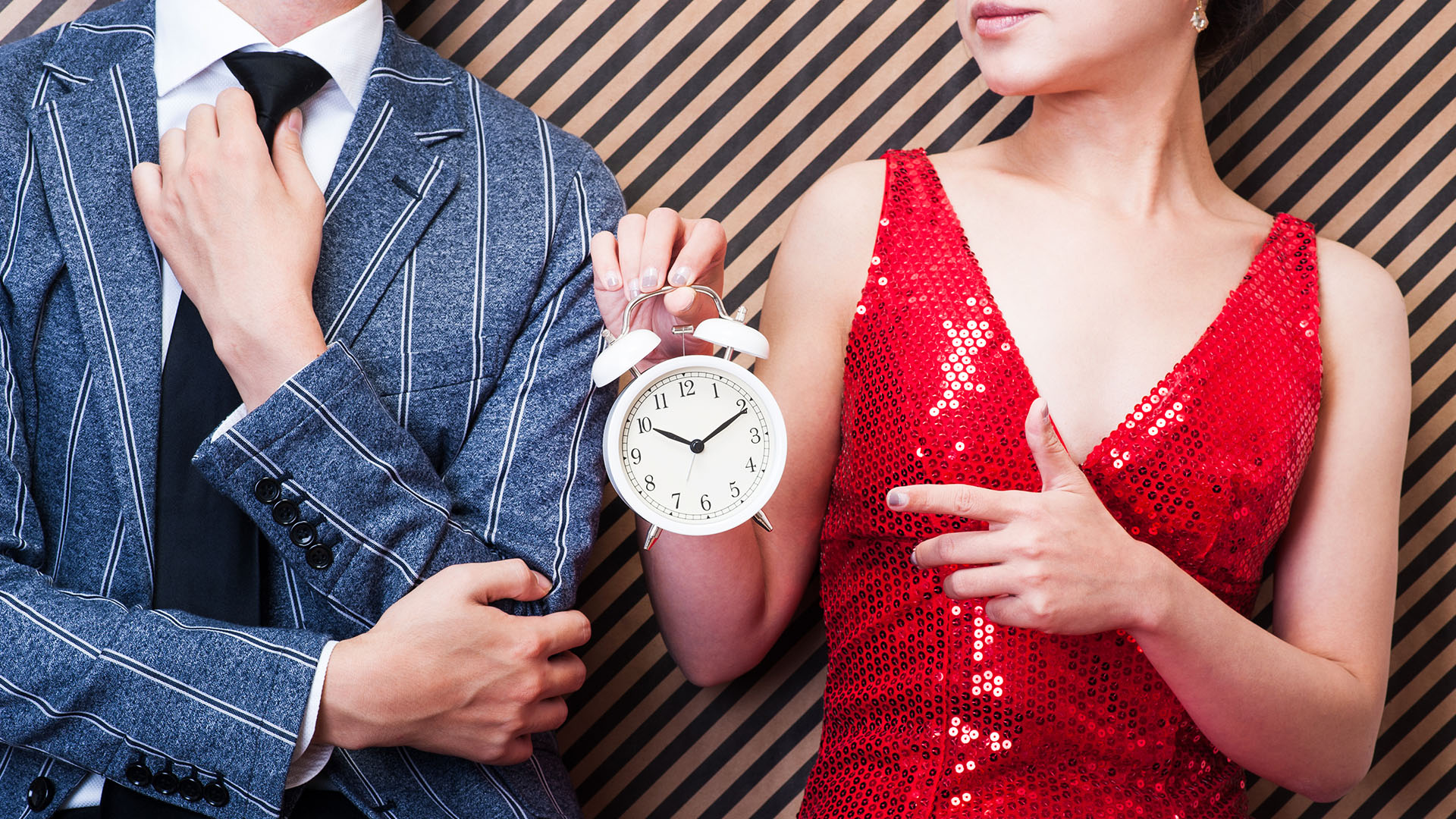 Body of a man in a suit adjusting his tie and body of a women in red shiny dress holding an alarm clock and pointing at it.