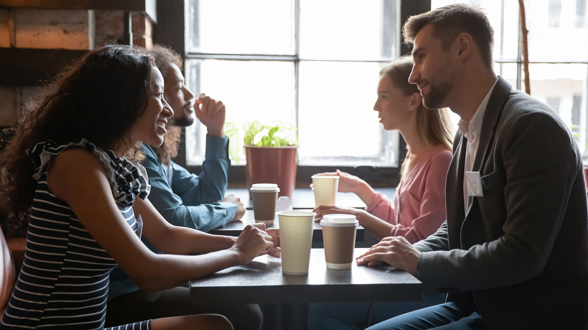 Two men and two women sitting next to each other at a cafe with coffees in front of them, conversing with each other.
