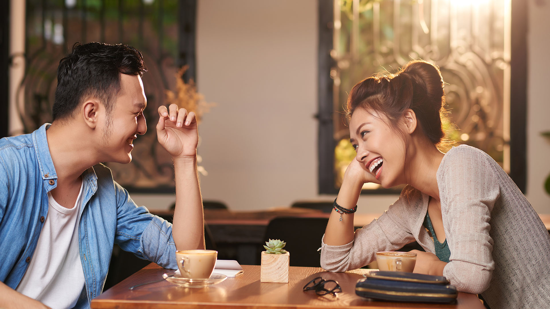 Dark-haired man and brunette woman laughing while looking at each other, they are sitting at a cafe having coffee.