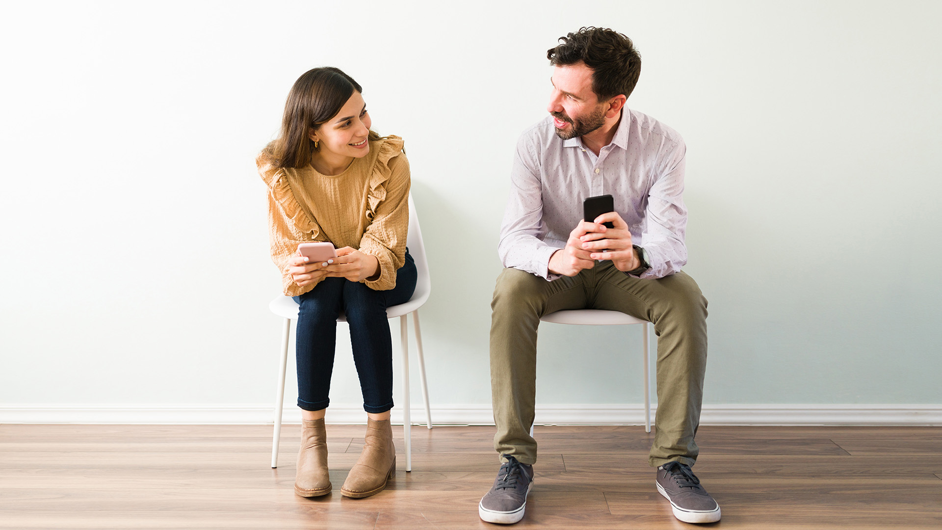 Brunette woman and dark-haired man sitting on chairs next to each other, looking at each other while holding their phones.