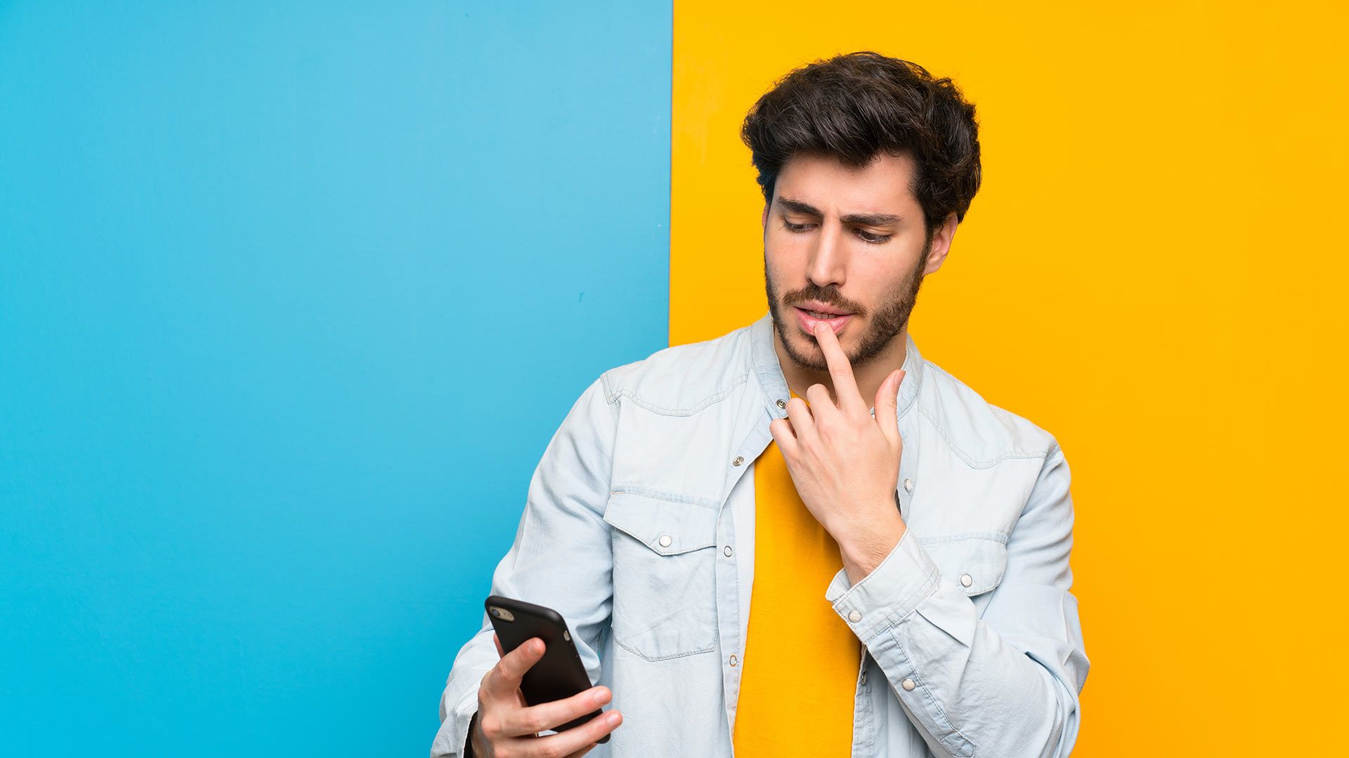 Brunette man wearing a yellow shirt looking at his phone and touching his lower lip. Split blue and yellow background.