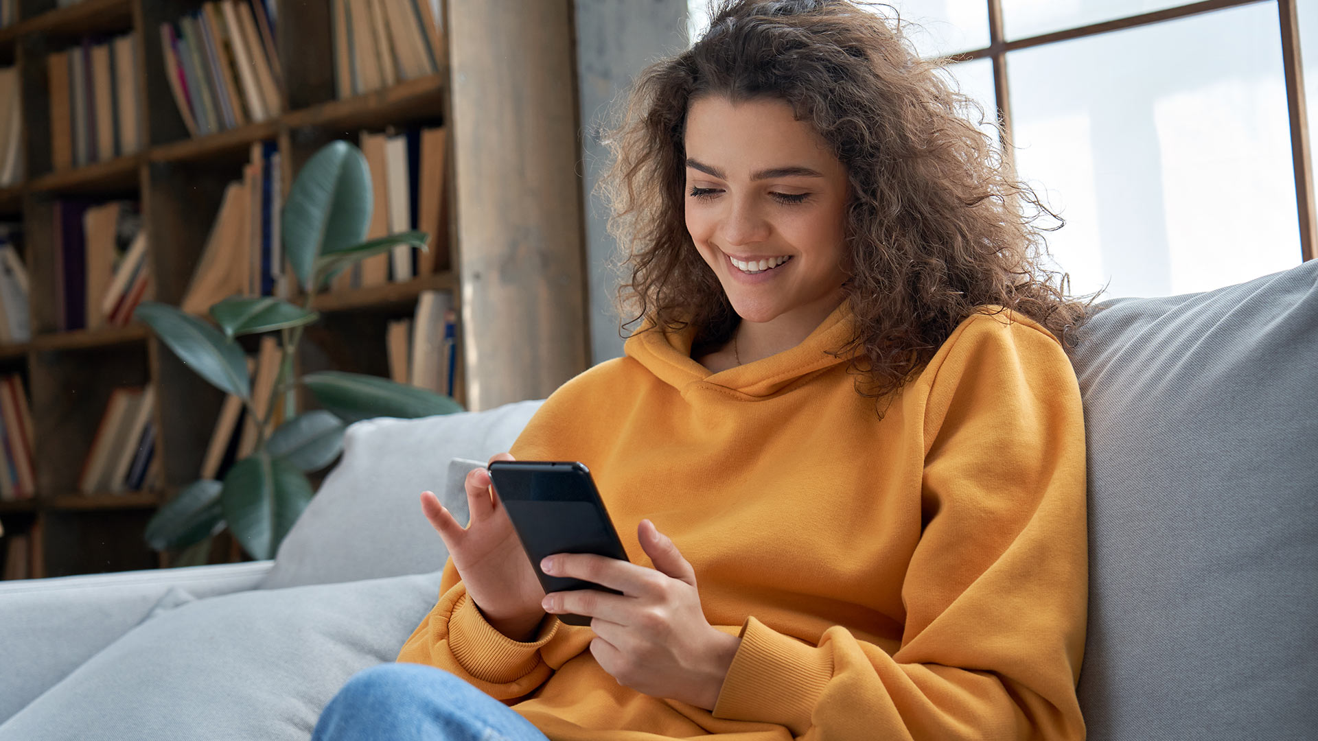 Brunette woman in yellow hoodie sitting down on a grey couch and looking at her phone while smiling.