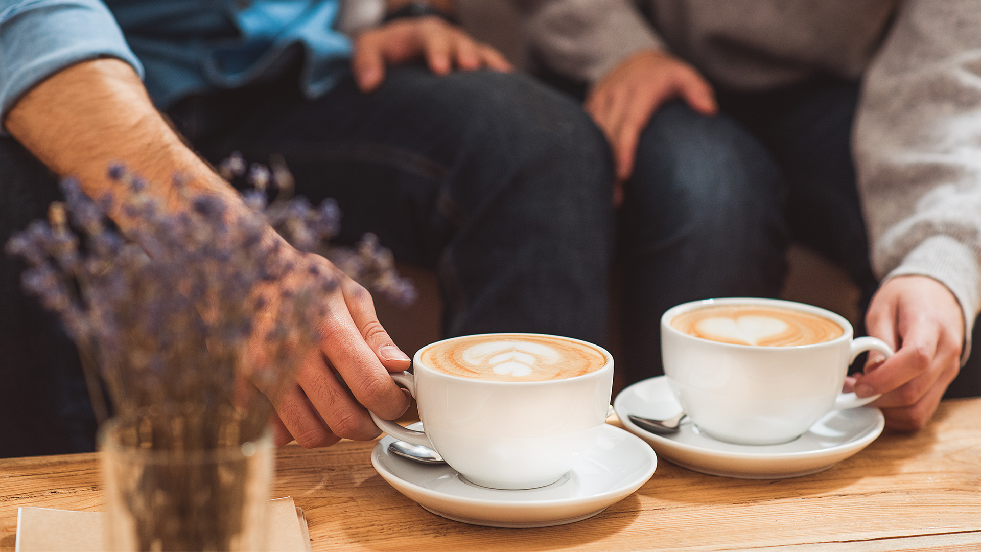 Hands of a man and woman sitting down at a cafe and grabbing their coffees. The coffee foam has love hearts drawn on it.